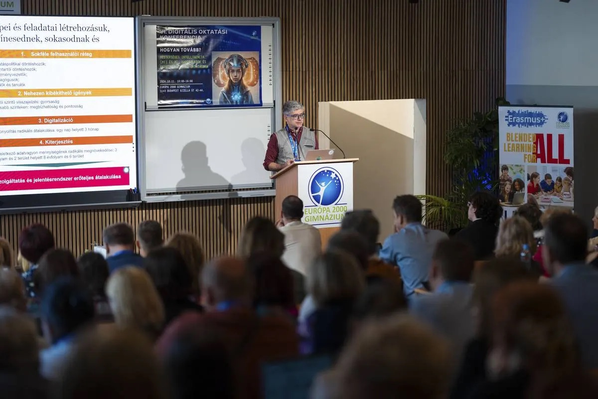 Előadás a VI. Digitális Oktatási Konferencián: előadó a pódiumon, mellette a nagy interaktív kijelző, előtérben a teltházas közönség
